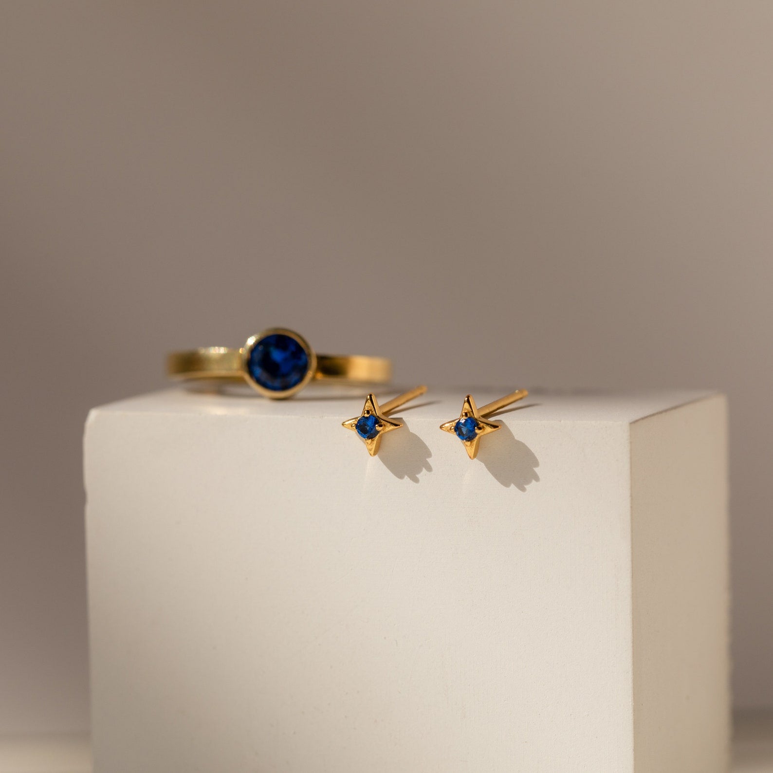 Sapphire Star Studs and a gold ring with a blue stone are elegantly displayed on a white block.