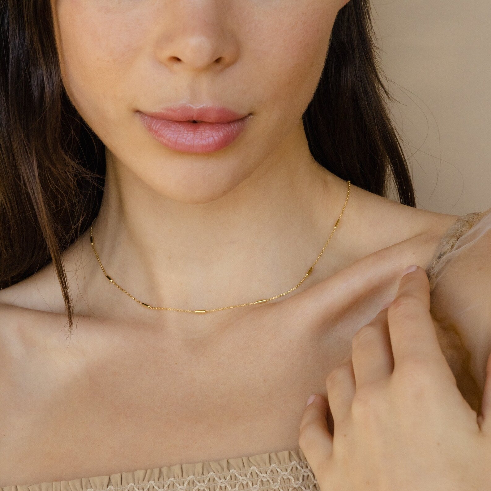A woman with natural makeup stands against a light neutral background, wearing the Bar Station Necklace—delicate chains gently draping and touching her collarbone.