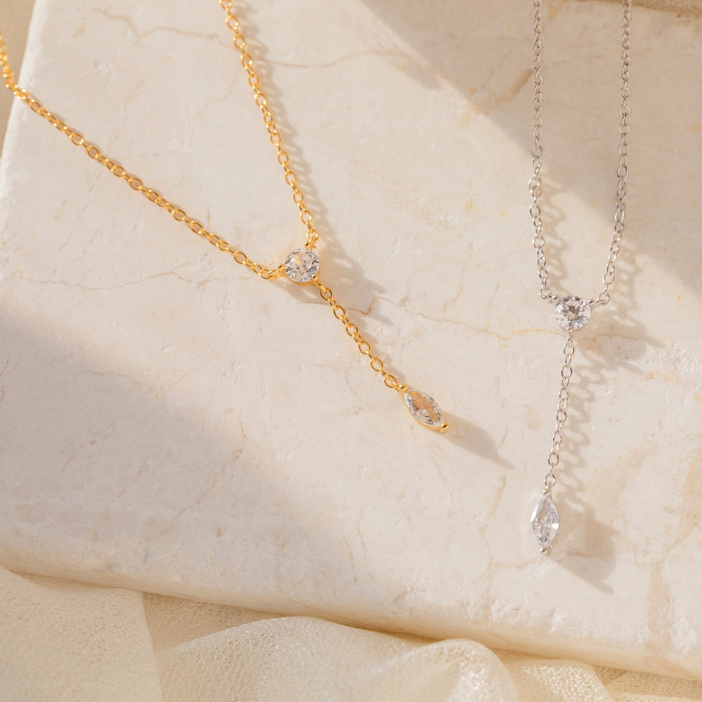 The Leena Diamond Lariat Necklace, featuring both round and marquise-shaped gemstones in a delicate lariat design, is displayed elegantly on a beige surface.