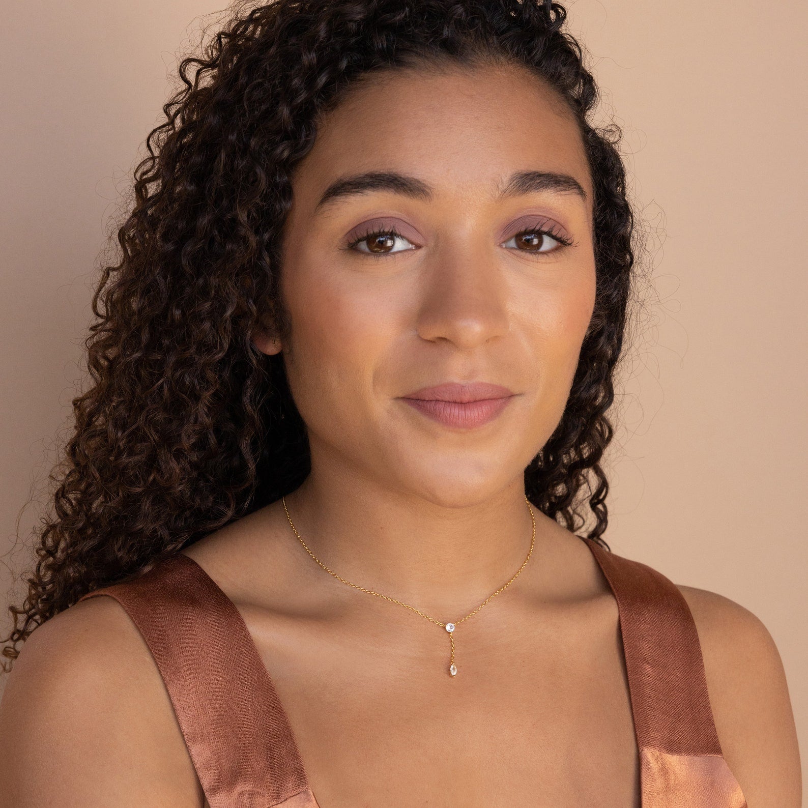 Woman with curly hair wearing a brown satin top and the Leena Diamond Lariat Necklace, featuring a delicate chain, posing against a beige background.