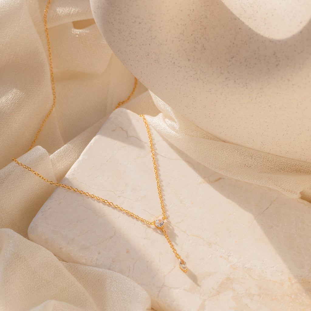 The Leena Diamond Lariat Necklace, a delicate gold chain with two small pendants, is displayed on a light marble surface with soft fabric.