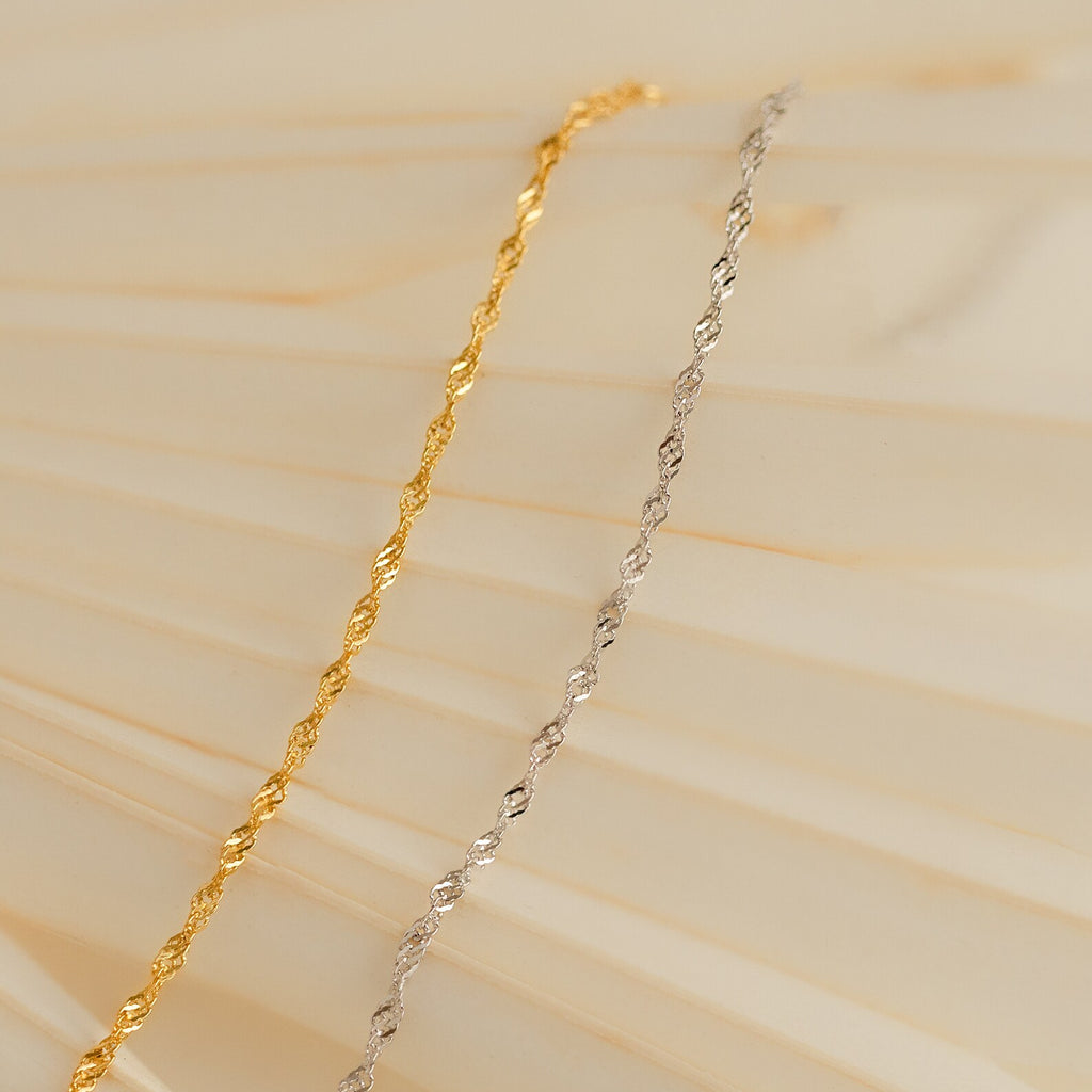 The Singapore Twist Bracelet, shown in gold and silver on a soft beige wavy background, highlights its elegant and minimalist style.
