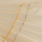 The Singapore Twist Bracelet, shown in gold and silver on a soft beige wavy background, highlights its elegant and minimalist style.