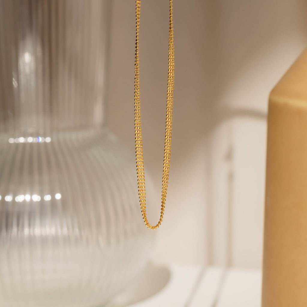 The Tri-Stacked Beaded Necklace, featuring a delicate gold chain and beads, is styled in front of a glass vase and beige backdrop—a versatile jewelry piece ideal for stacking and suitable for any occasion.