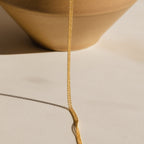The Tri-Stacked Beaded Necklace, a delicate gold chain ideal for layering with minimalist pieces, is displayed draped over the edge of a tan ceramic bowl on a beige surface.
