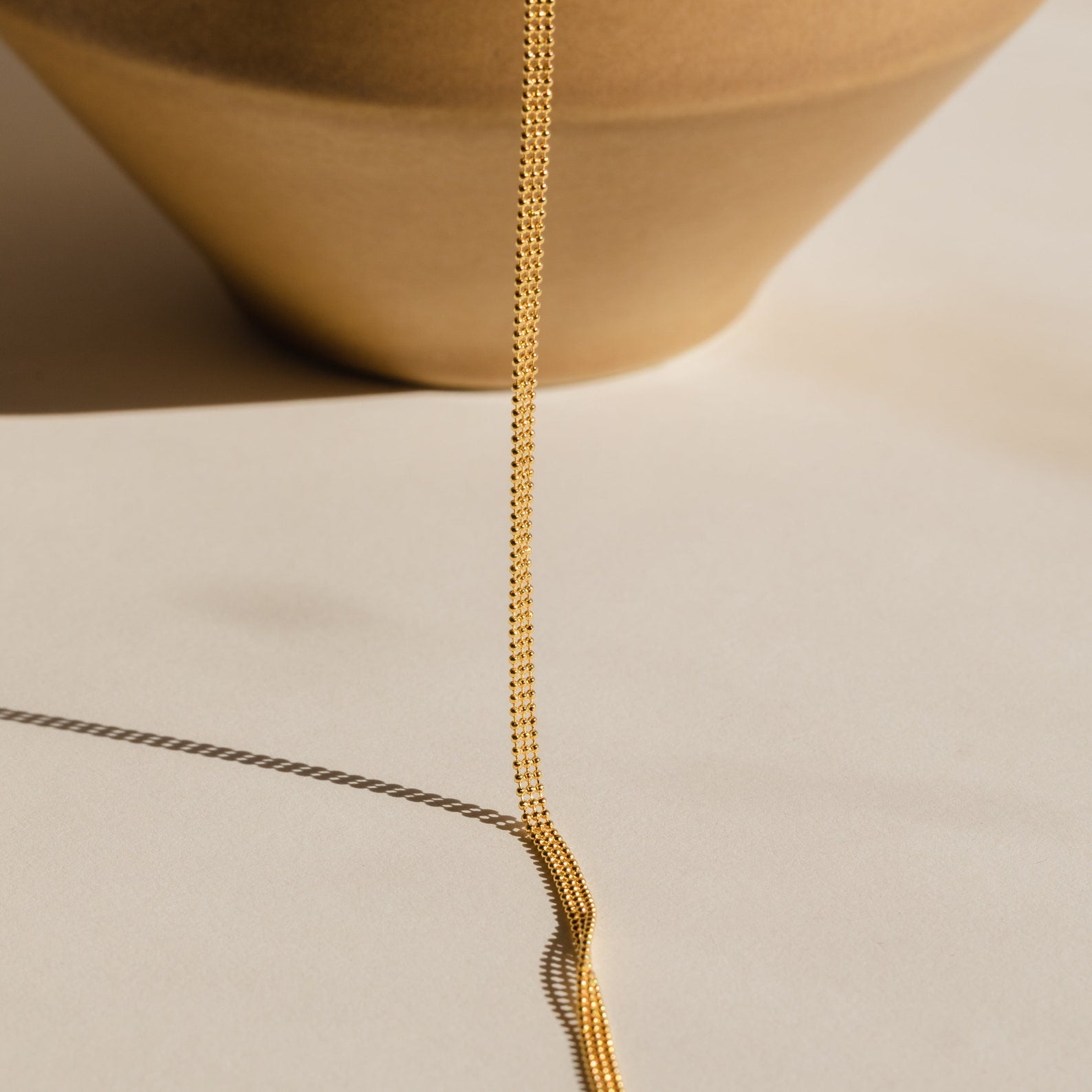 The Tri-Stacked Beaded Necklace, a delicate gold chain ideal for layering with minimalist pieces, is displayed draped over the edge of a tan ceramic bowl on a beige surface.