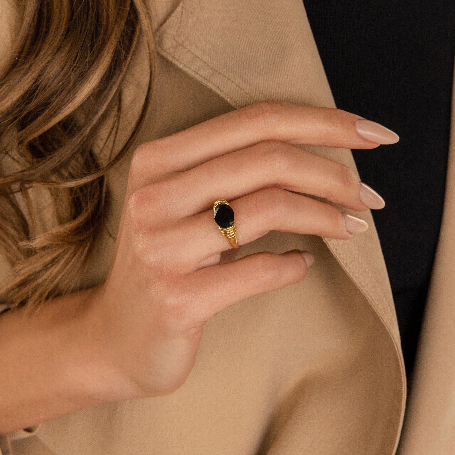A woman's hand with nude nails wears the Dusk Signet Ring—vintage-inspired with a black stone—set against a beige blazer, showcasing timeless statement jewelry.