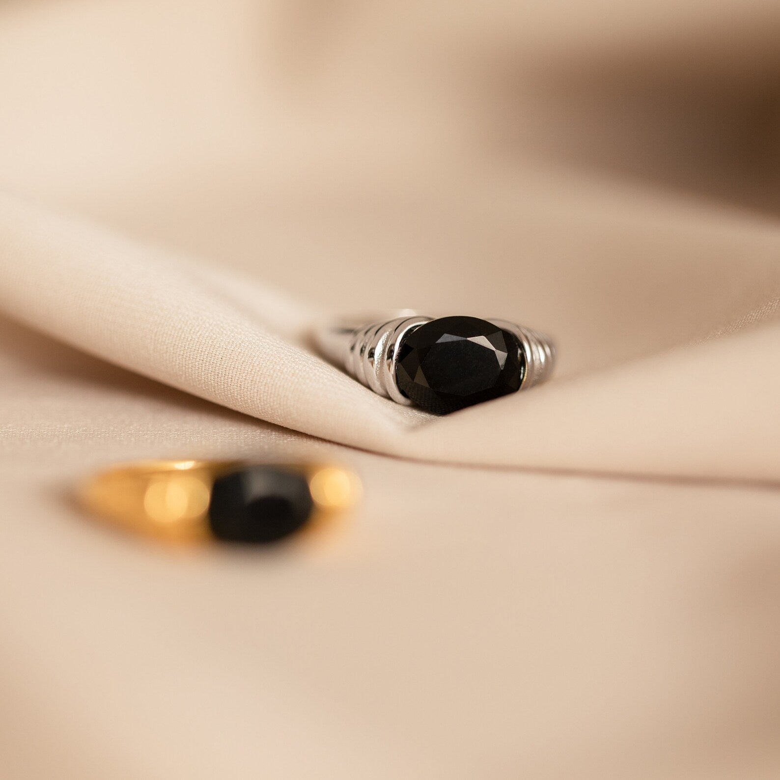 The Dusk Signet Ring, featuring a striking black gemstone, rests on beige fabric with a blurred gold ring in the foreground—ideal for anyone who appreciates bold statement jewelry.