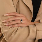 A hand with nude nails wears the Dusk Signet Ring, featuring a black stone, resting on a tan trench coat sleeve—a subtle nod to statement jewelry.