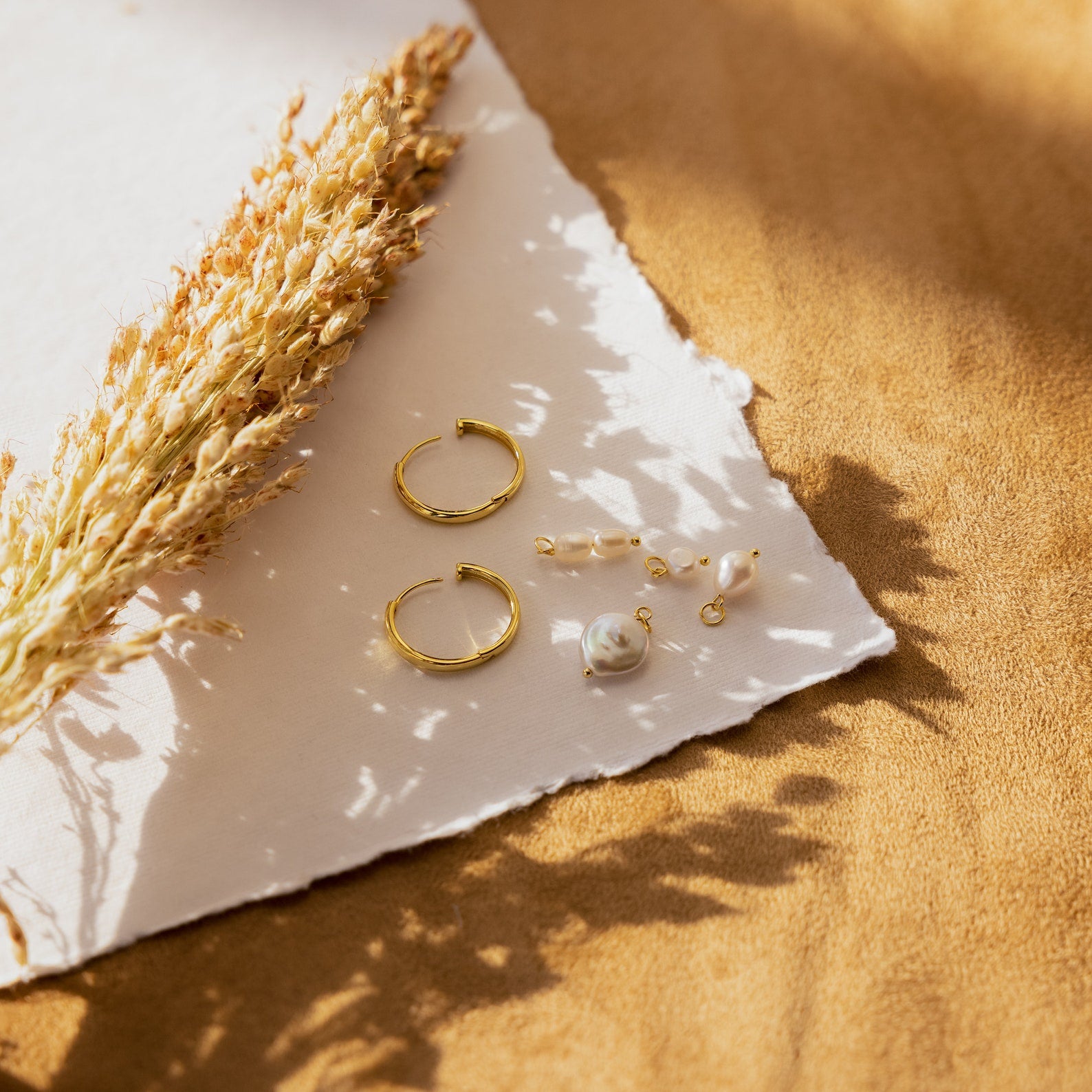 The Pearl Drop Earrings Set rests on white fabric beside dried grass. Warm sunlight and soft shadows highlight these minimalist bridal earrings, perfect for adding elegance to any special occasion.