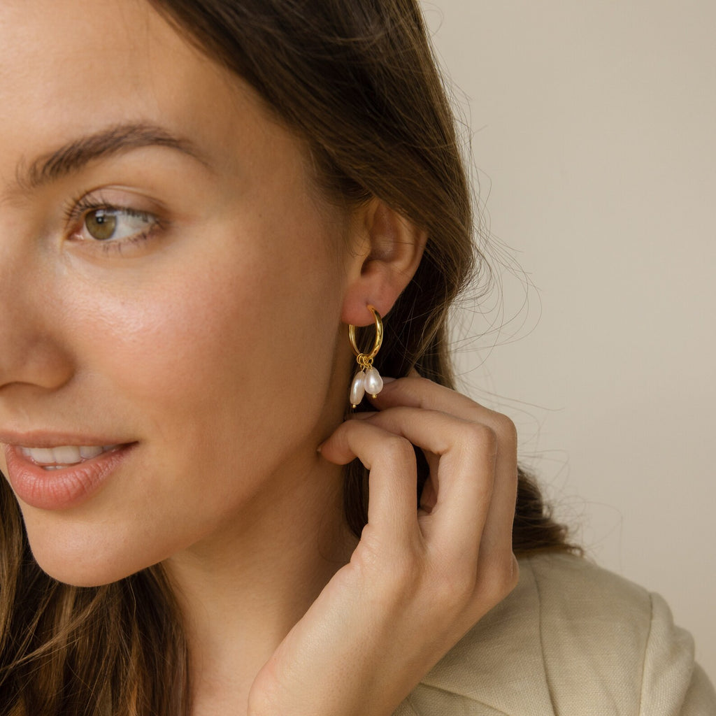 A woman with long brown hair smiles softly as she touches her Pearl Drop Earrings Set featuring elegant pearl charms.