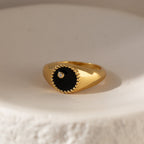 The Natasha Black Signet Ring features a round black onyx face, a central CZ diamond, and a twisted gold border, displayed on a white surface.