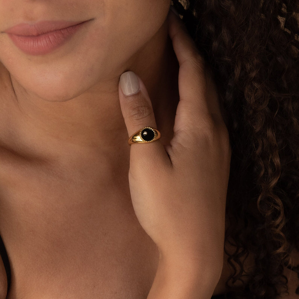 A woman touches her neck, wearing the Natasha Black Signet Ring—a gold band set with a striking black stone—on her finger.
