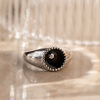 The Natasha Black Signet Ring features a sleek silver band with a black circular top and a small CZ diamond at the center, displayed on a marble surface.