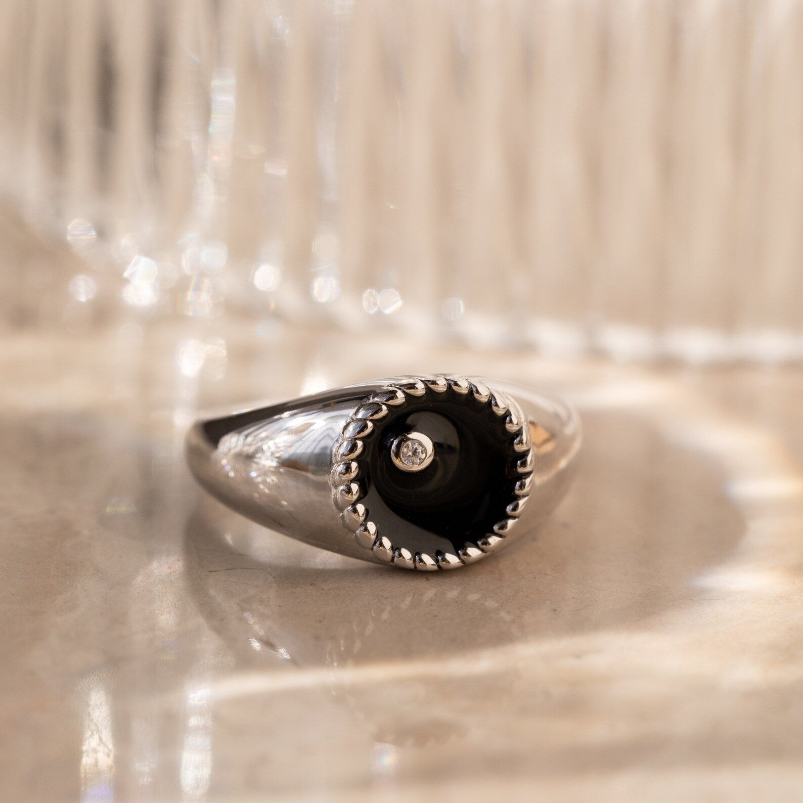 The Natasha Black Signet Ring features a sleek silver band with a black circular top and a small CZ diamond at the center, displayed on a marble surface.