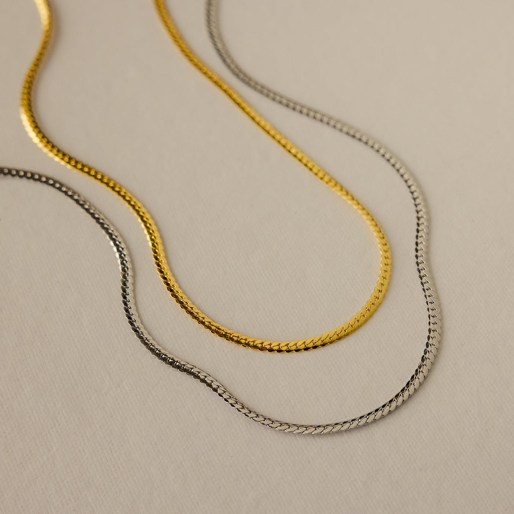 Gold and silver herringbone chain necklaces with a sleek, flat design, displayed side by side on a neutral background.