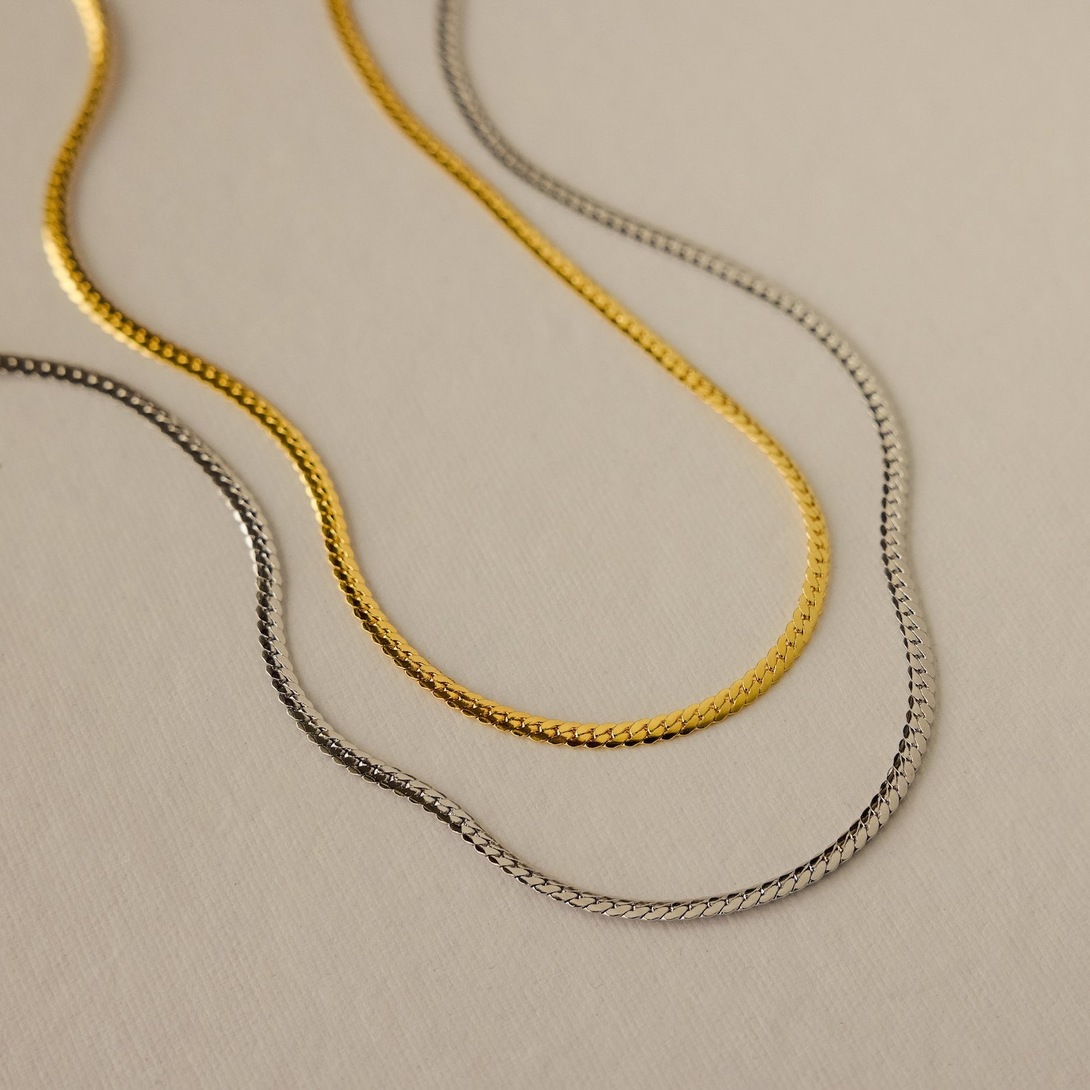 Gold and silver herringbone chain necklaces with a sleek, flat design, displayed side by side on a neutral background.