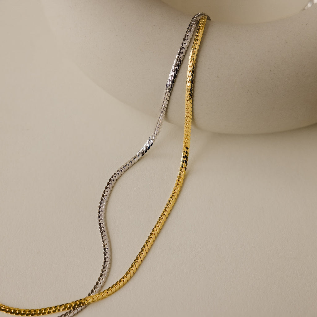 The Thick Snake Chain Necklace, shown draped over a smooth, round, off-white ceramic piece on a beige background, is an effortless everyday staple.