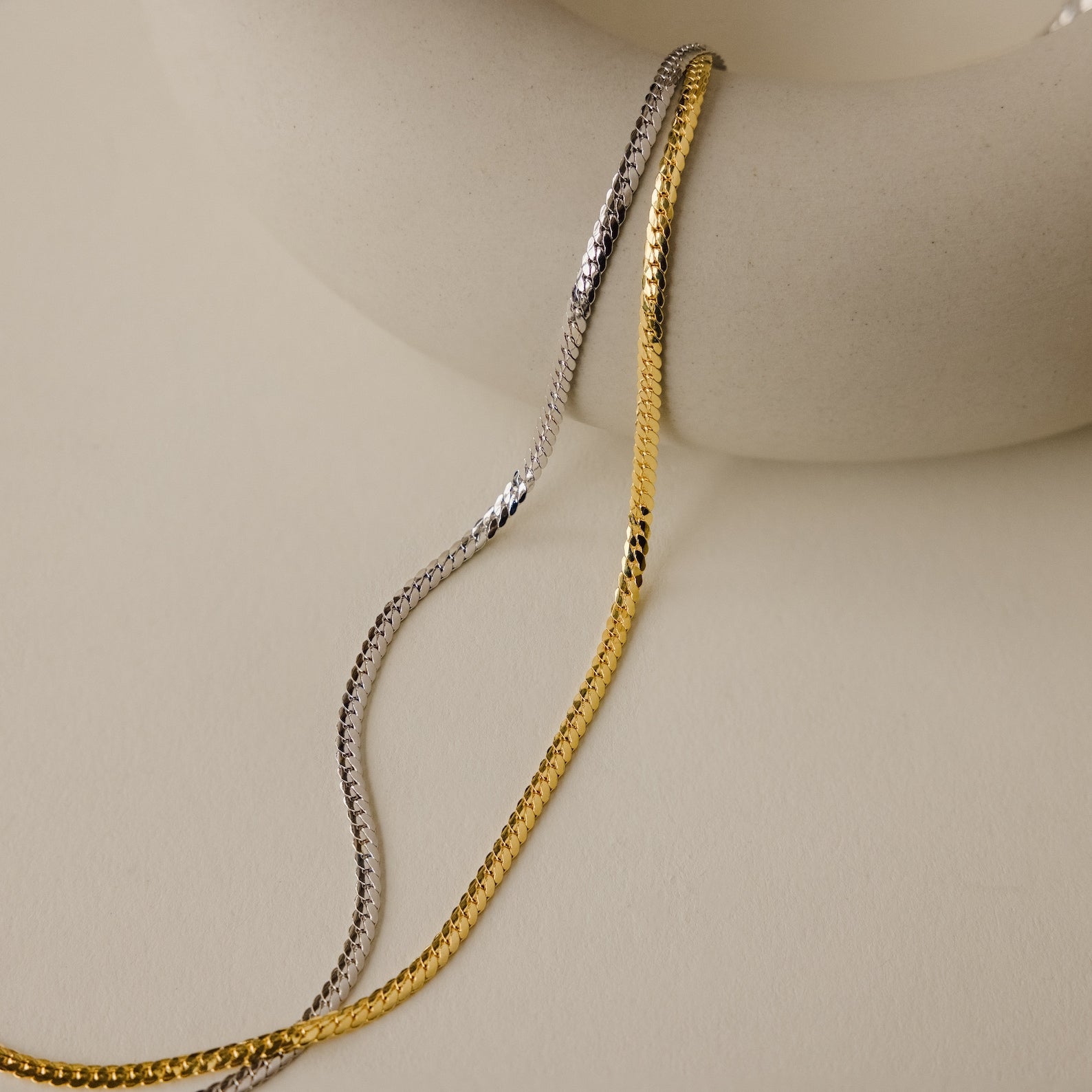 The Thick Snake Chain Necklace, shown draped over a smooth, round, off-white ceramic piece on a beige background, is an effortless everyday staple.