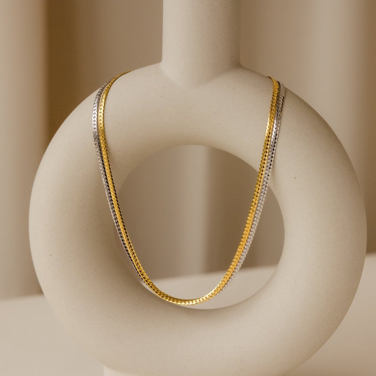 The Thick Snake Chain Necklace, in gold and silver, draped over a modern round beige ceramic vase—an effortless everyday staple.