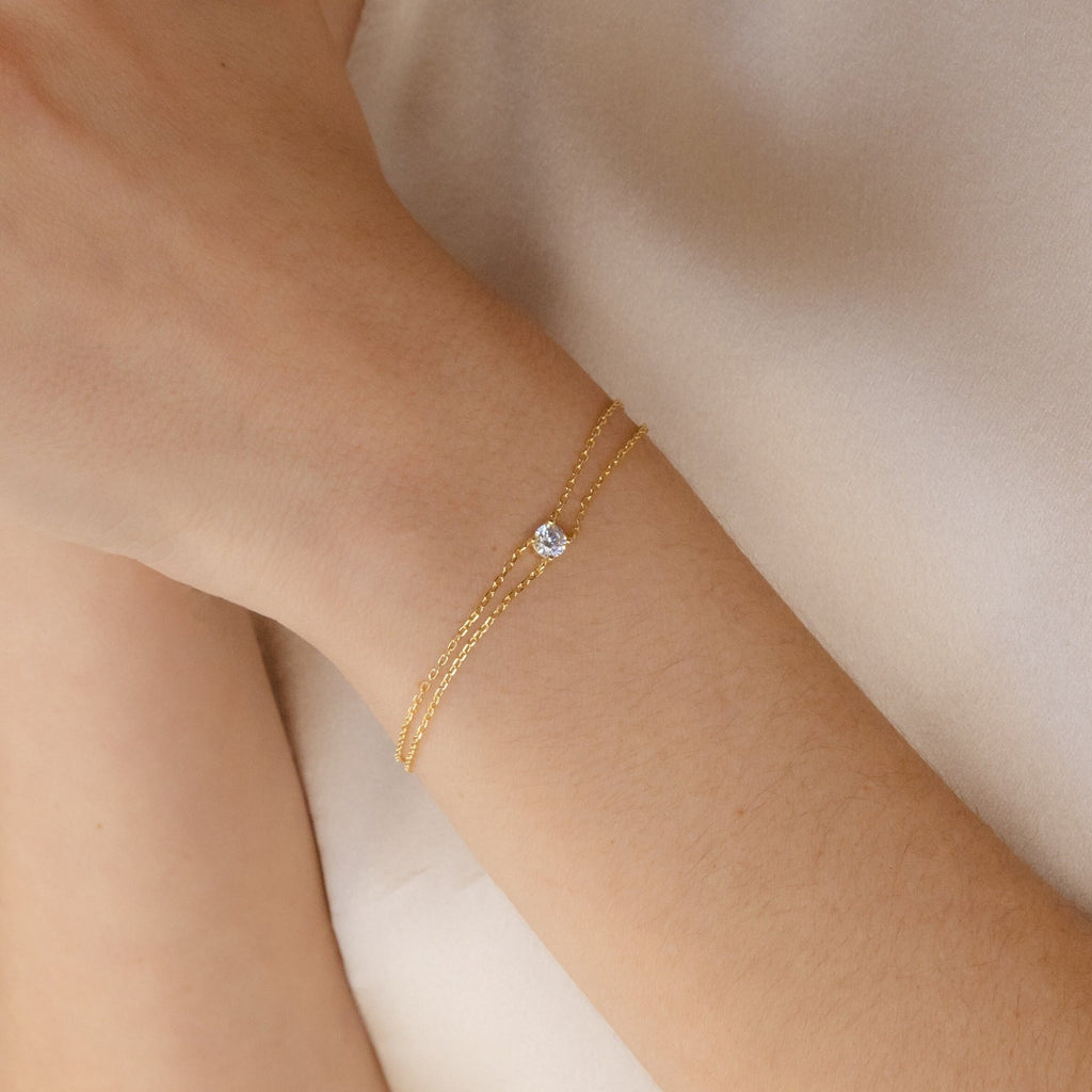 The Duo Chain Diamond Bracelet, a minimalist gold chain adorned with a single diamond, rests gracefully on a wrist against light fabric—a refined and elegant anniversary gift choice.