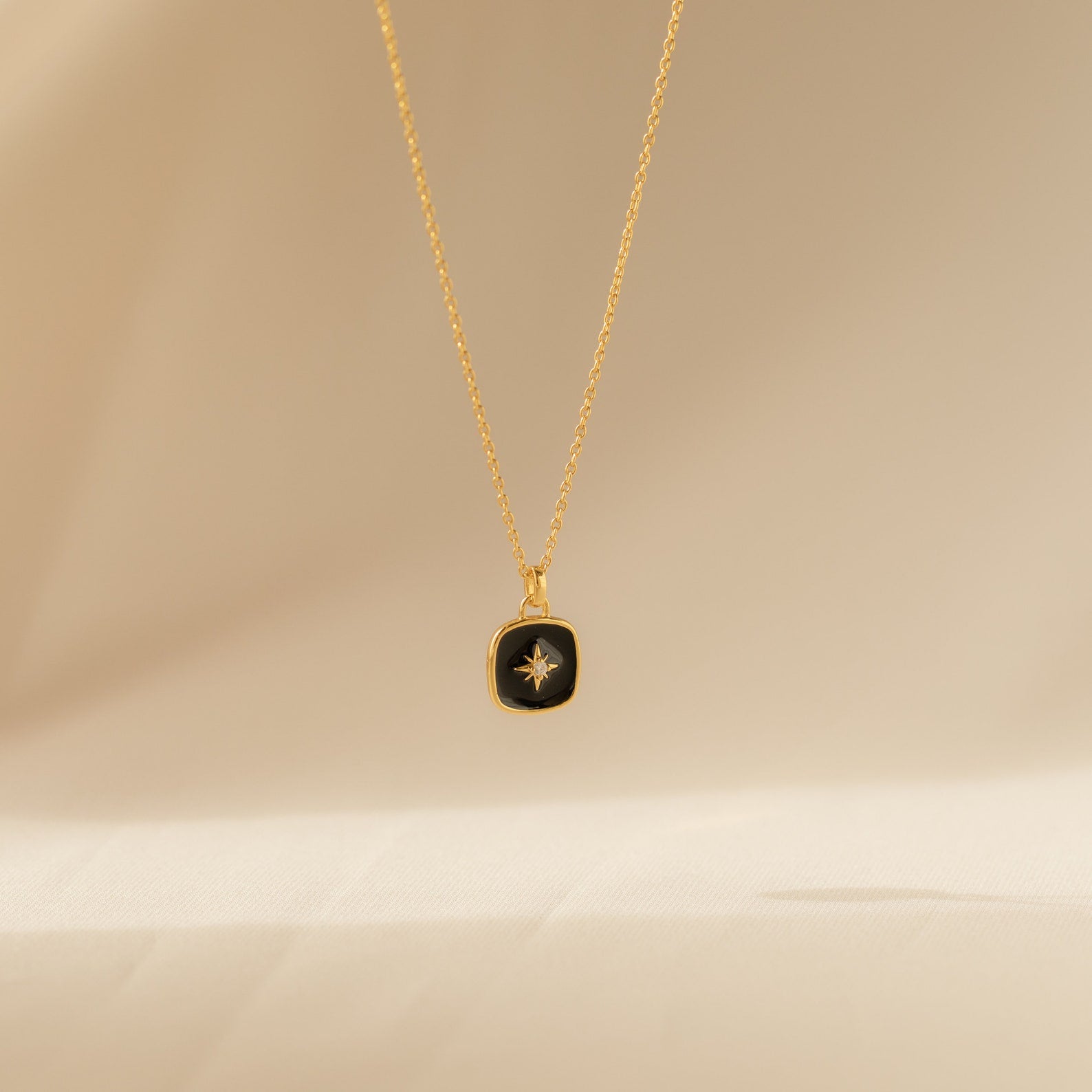 Hermoine Pendant Necklace with a gold chain and black pendant accented by a small star design, shown on a beige background.
