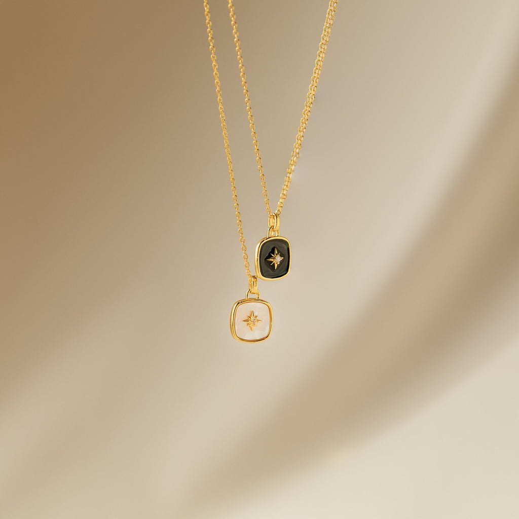 The Hermoine Pendant Necklace features two gold chains with square pendants—one black, one white—set against a neutral background, ideal for layering or pairing with your favorite Mother of Pearl piece.