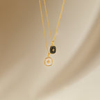 The Hermoine Pendant Necklace features two gold chains with square pendants—one black, one white—set against a neutral background, ideal for layering or pairing with your favorite Mother of Pearl piece.