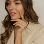 A woman with wavy brown hair in a beige blazer touches her face, highlighting the Crescent Pearl Ring—a delicate handmade stacking ring ideal for minimalist jewelry lovers.