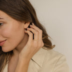 A woman with long brown hair smiles softly, touching her ear while wearing the Crescent Pearl Ring and a beige collared shirt.