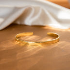 The Alyssa Cuff Bracelet, featuring an elegant gold and silver cuff design, is displayed on a brown surface with a white cloth in the background.