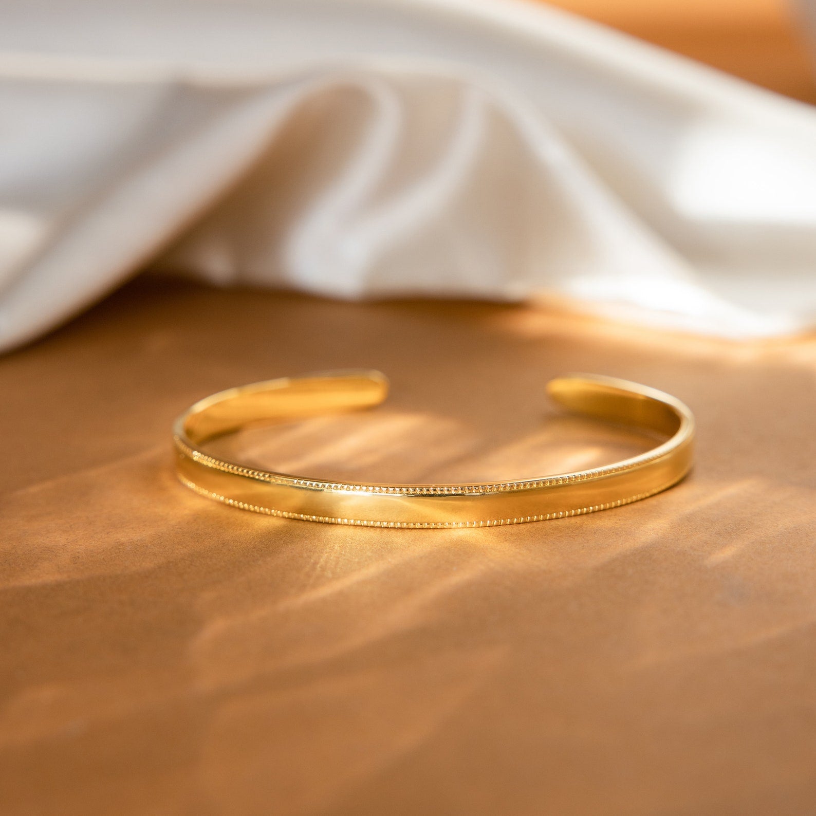 The Alyssa Cuff Bracelet, featuring an elegant gold and silver cuff design, is displayed on a brown surface with a white cloth in the background.