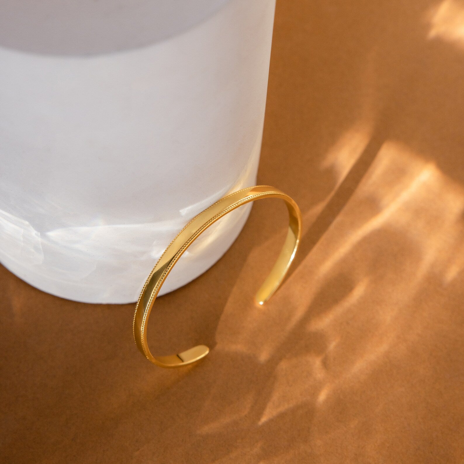 The Alyssa Cuff Bracelet in gold is displayed on a white cylindrical stand atop a brown surface, illuminated by soft light and gentle shadows.