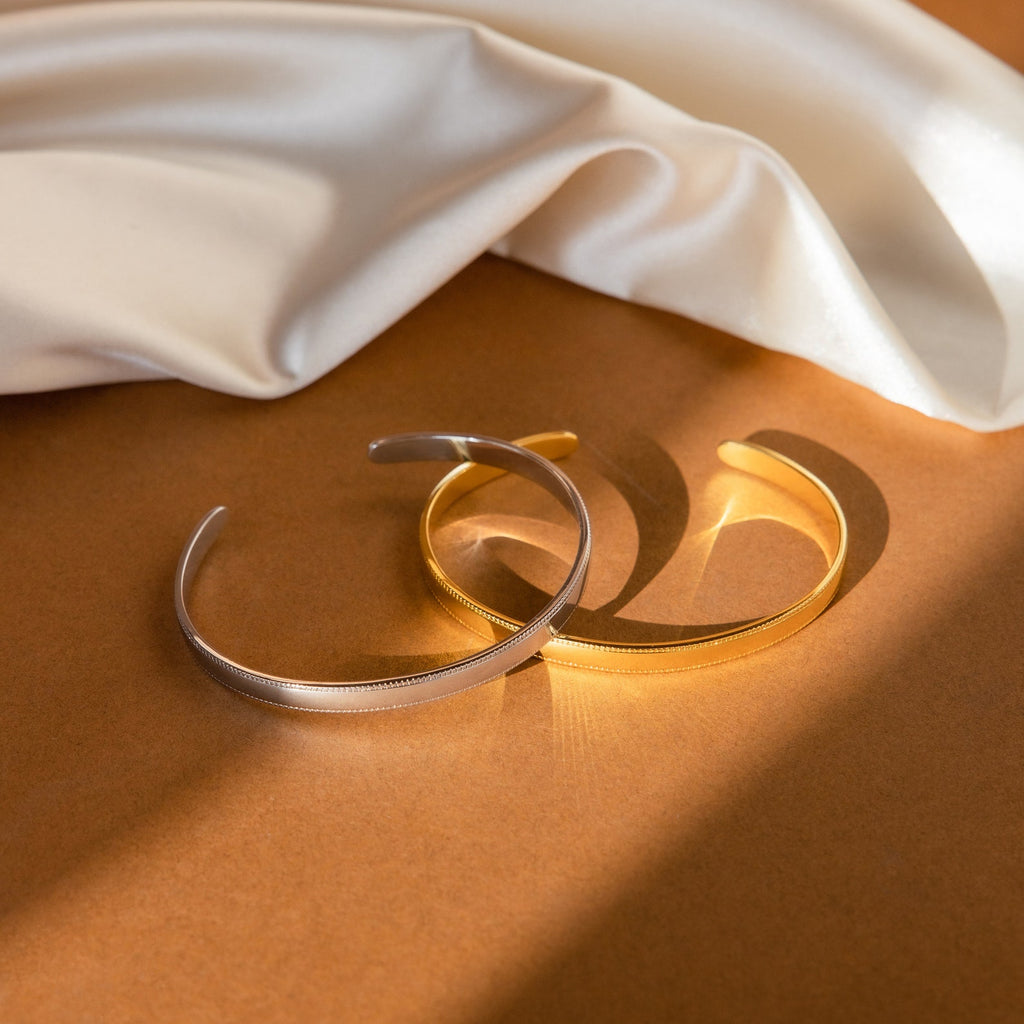Two metallic Alyssa Cuff Bracelets, one in gold and one in silver, rest on a brown surface beside draped white fabric.