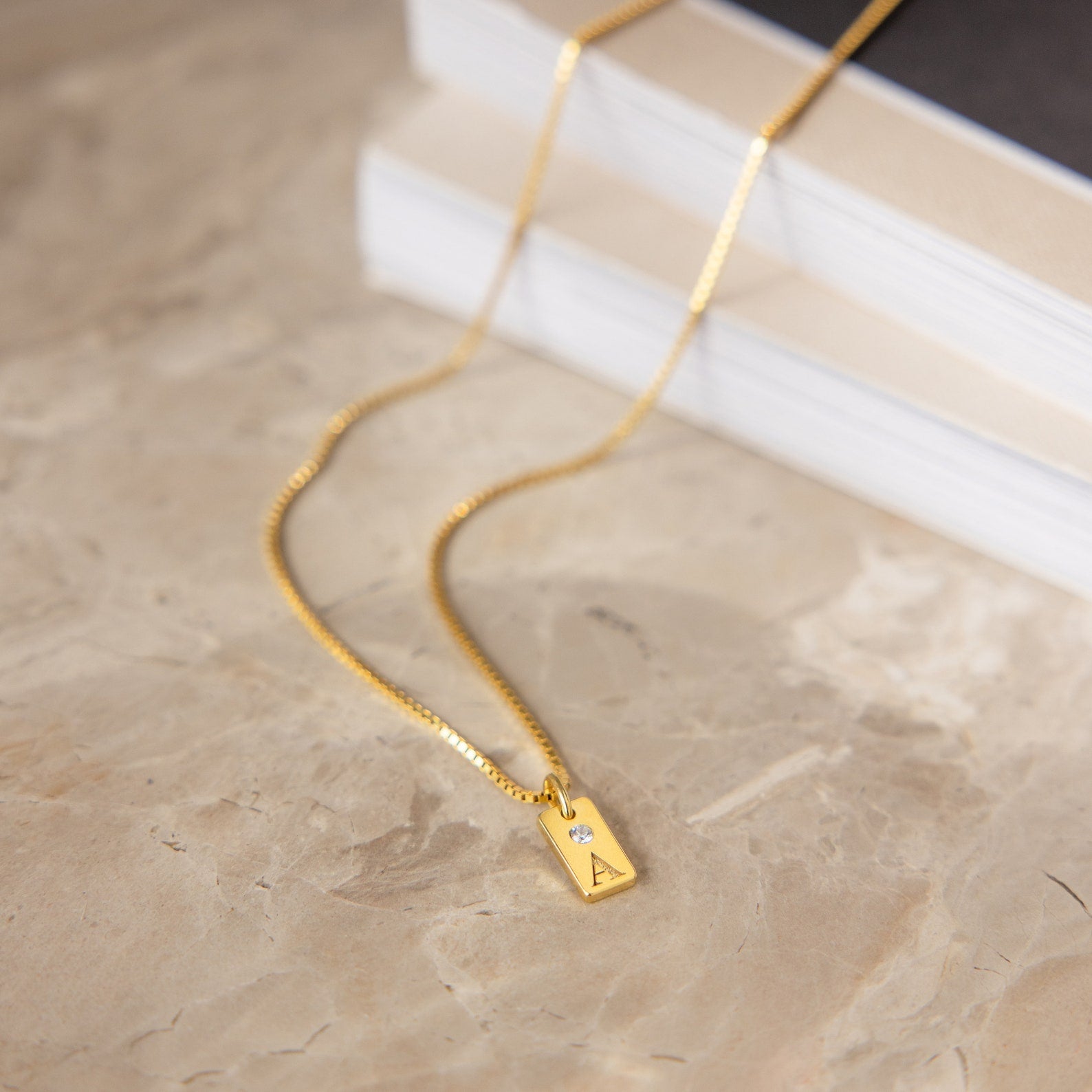 The Mini Birthstone Tag Necklace showcases a gold rectangular pendant engraved with the letter A and accented by a small gemstone, displayed on a marble surface.
