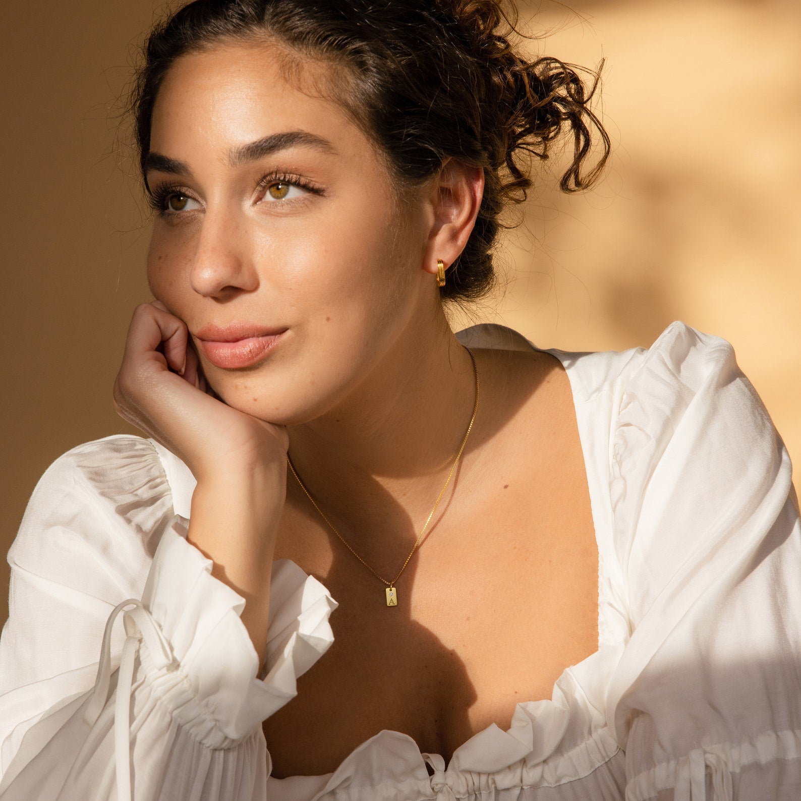 A woman in a white blouse rests her chin on her hand, wearing gold jewelry, including the Mini Birthstone Tag Necklace, and gazes thoughtfully to the side.