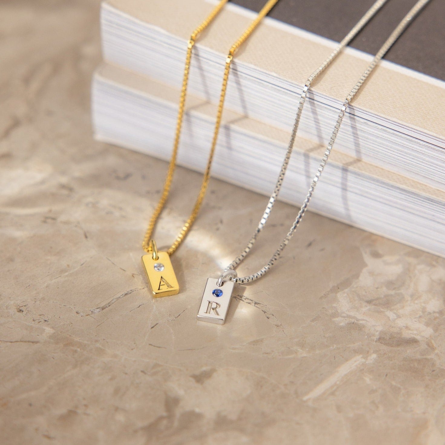 The Mini Birthstone Tag Necklace, personalized with letter A and R pendants in gold and silver, is displayed on a marble surface next to stacked books.