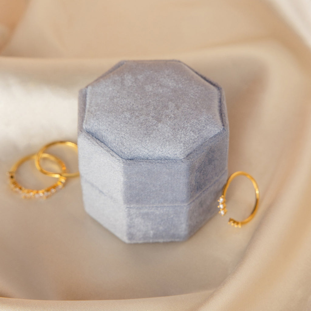 The 3 Slot Velvet Ring Box in gray, featuring a hexagonal shape, is placed beside two gold rings on a soft beige fabric background.
