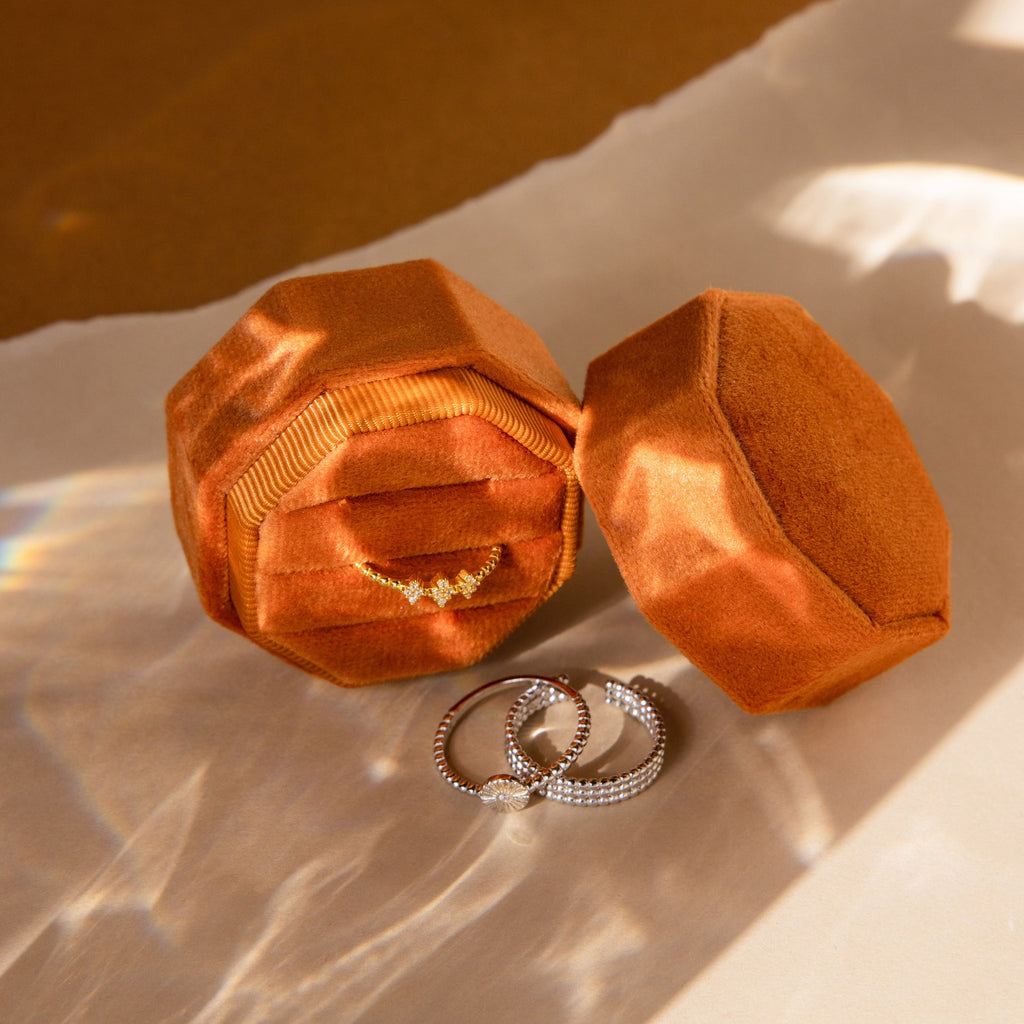 The 3 Slot Velvet Ring Box in orange holds a diamond ring inside, with two rings displayed in front on a sunlit surface—a chic organizer for your jewelry collection.