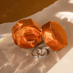 The 3 Slot Velvet Ring Box in orange holds a diamond ring inside, with two rings displayed in front on a sunlit surface—a chic organizer for your jewelry collection.
