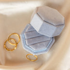 The 3 Slot Velvet Ring Box in light blue is open beside three gold rings on cream-colored fabric, creating an elegant jewelry display.