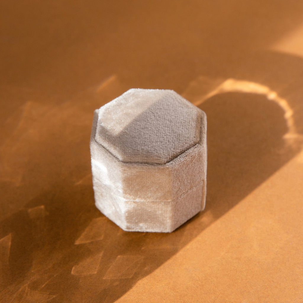 A 3 Slot Velvet Ring Box in beige with a hexagonal design rests on a brown surface, its shape casting a clear shadow in the sunlight.