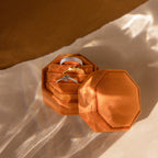 The Mystery Ring Box in orange velvet holds two silver rings and one gold ring, displayed on a beige surface with soft shadows.