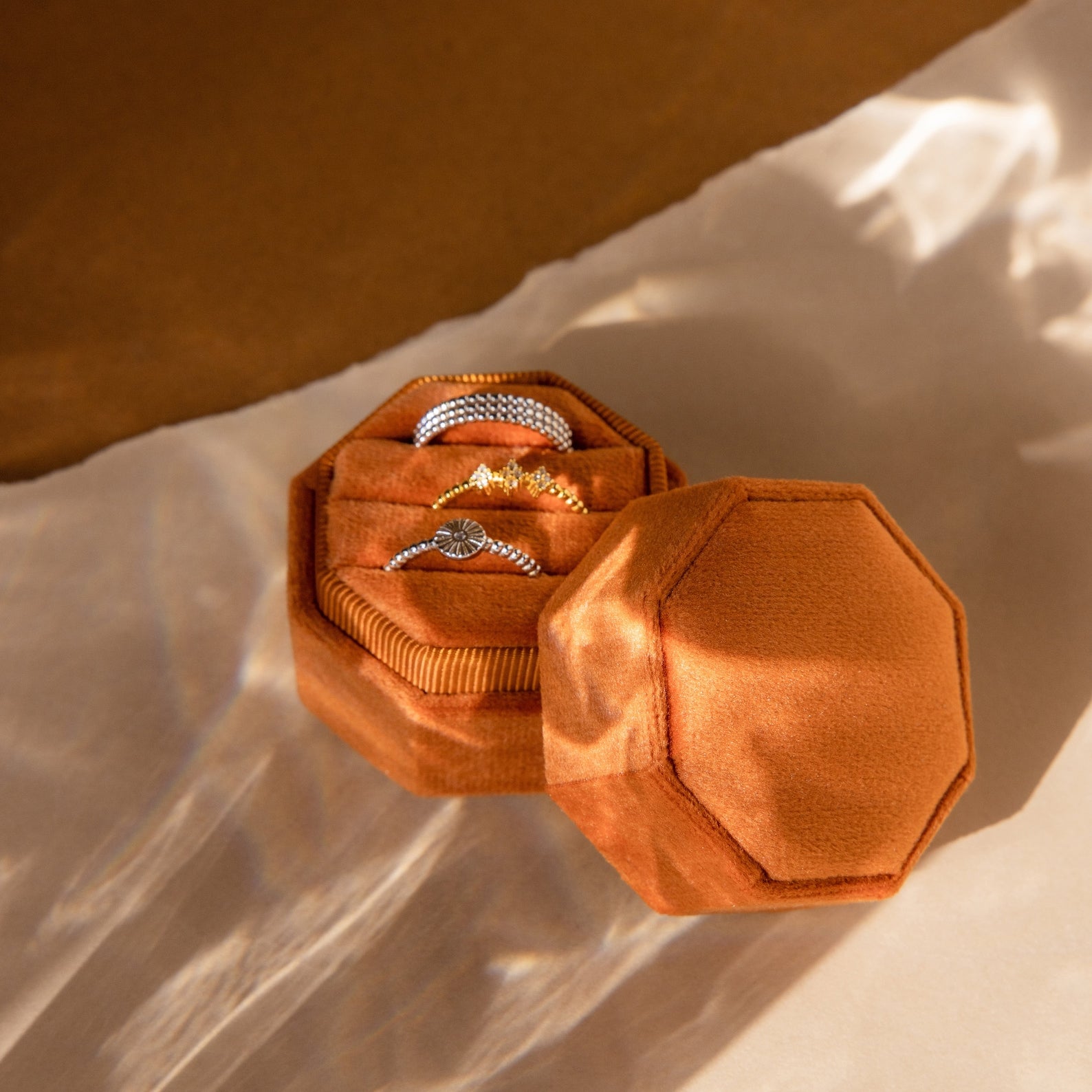 The Mystery Ring Box in orange velvet holds two silver rings and one gold ring, displayed on a beige surface with soft shadows.
