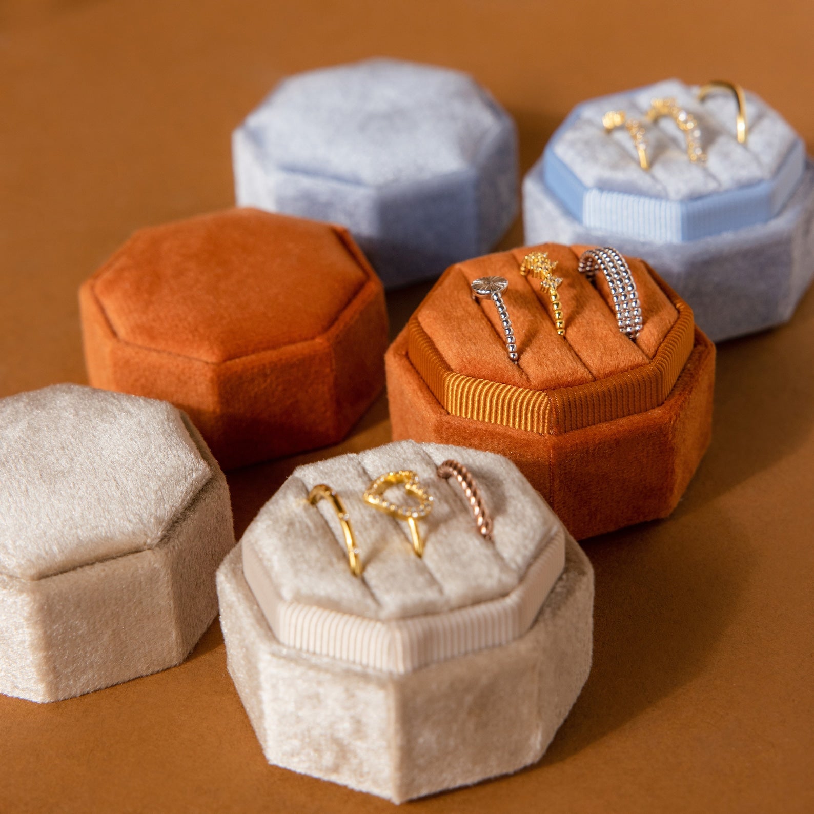 The Mystery Ring Box, available in tan, orange, and blue velvet, showcases gold and silver rings on a brown surface—each case adds elegance to your jewelry collection.