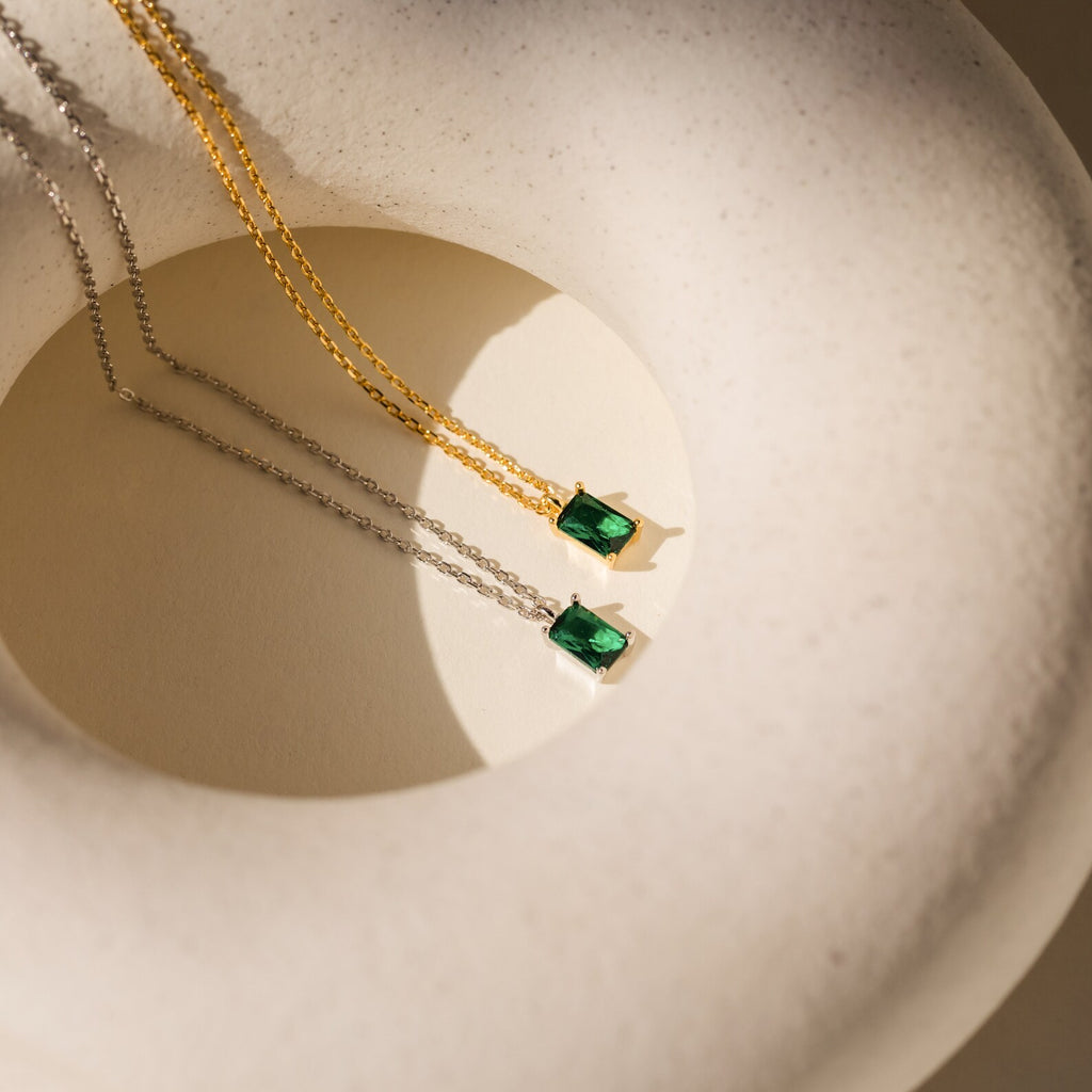 Two Maia Emerald Necklaces, one in gold and one in silver, each featuring a green emerald pendant, are displayed on a white textured surface.