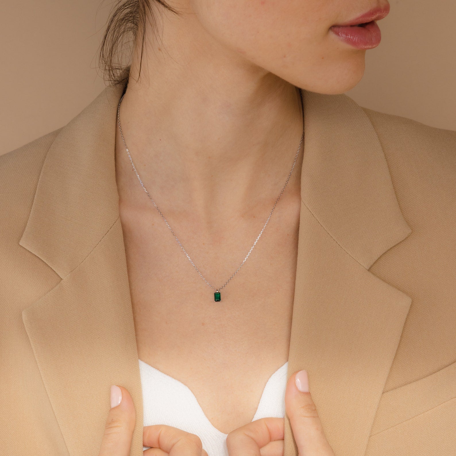 Woman in a beige blazer and white top showcases the Maia Emerald Necklace, a delicate silver necklace featuring a green gemstone pendant.