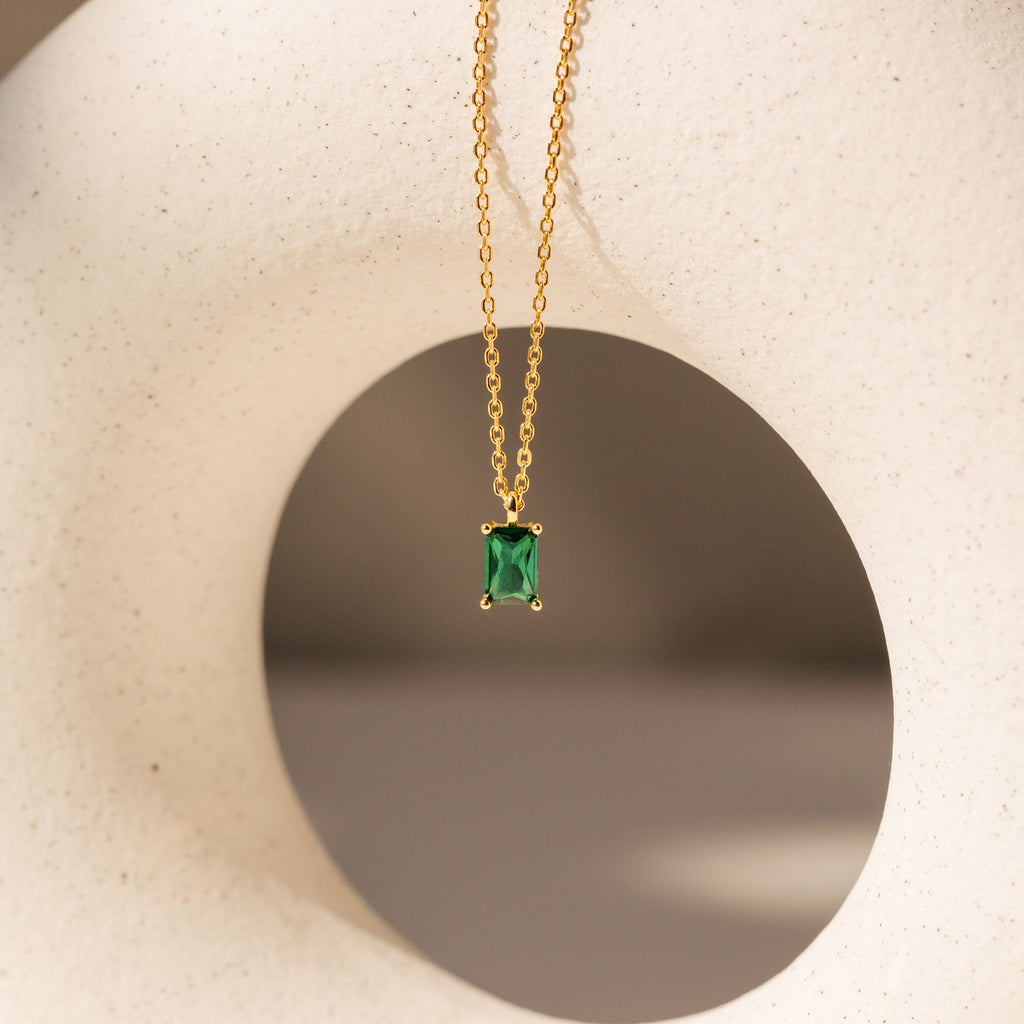 Maia Emerald Necklace featuring a gold chain and rectangular green emerald pendant, elegantly displayed against a white abstract sculpture.