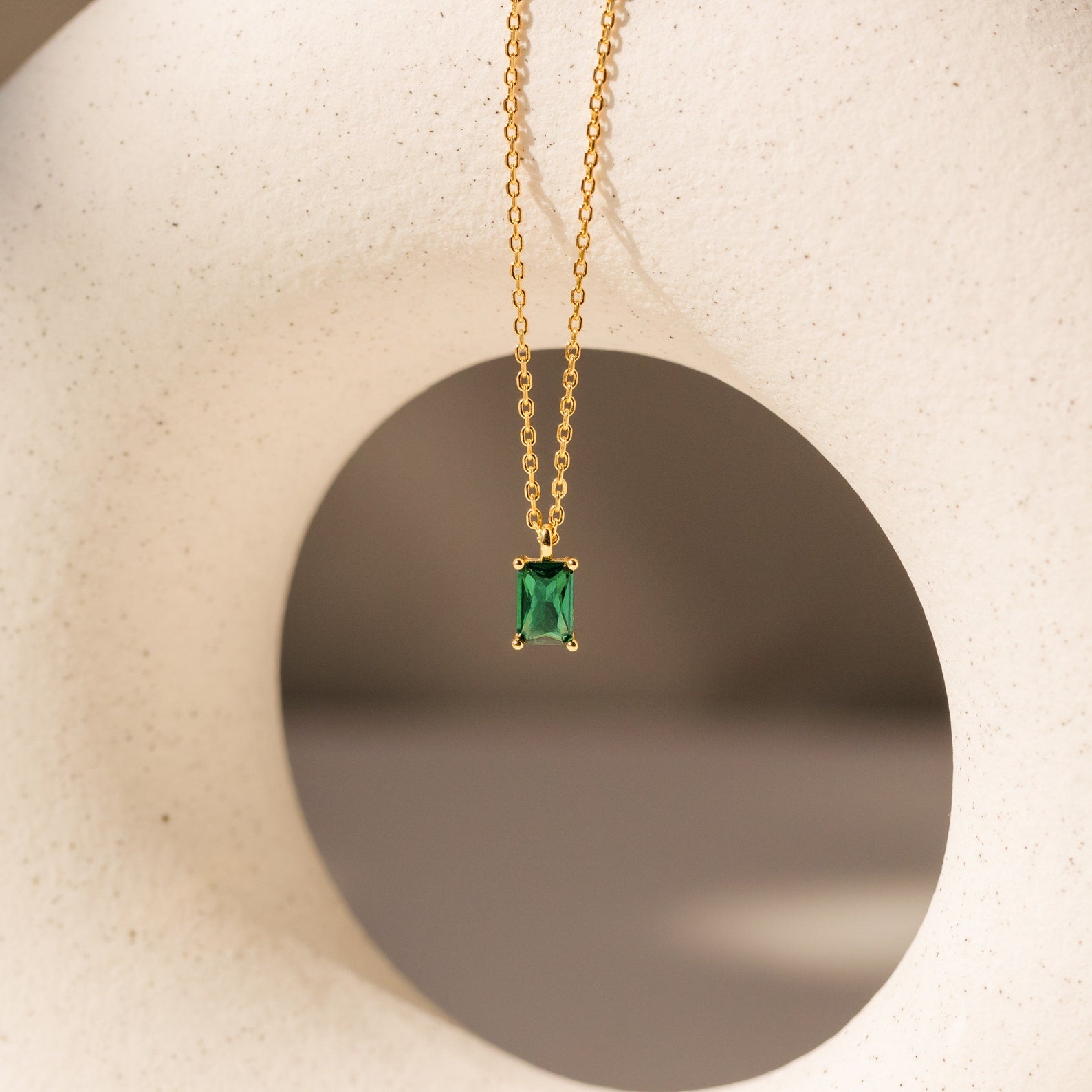 Maia Emerald Necklace featuring a gold chain and rectangular green emerald pendant, elegantly displayed against a white abstract sculpture.