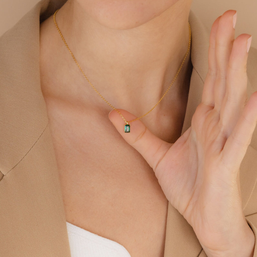 Someone wearing the Maia Emerald Necklace, a gold pendant featuring a small green emerald, gently holds it near their neck.